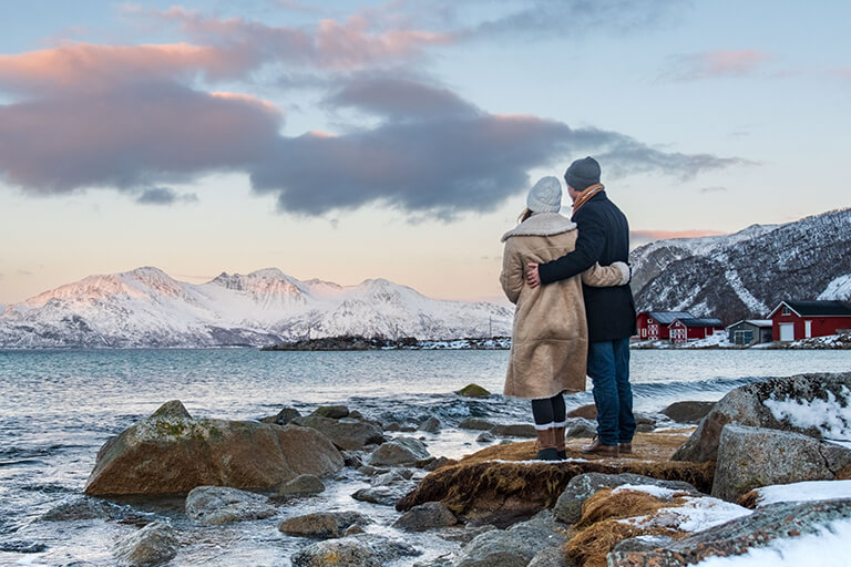 A couple dressed in winter coats and hats stand on a rocky shore, embracing as they look out over a snowy mountain landscape and calm water during sunset—perfect for Arctic adventures Norway. Red houses dot the background.