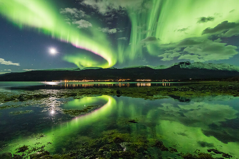 Bright green aurora borealis lights swirl over mountains near Tromso, reflecting on a calm lake with grassy, rocky shores. A crescent moon and distant town lights hint at unforgettable Arctic adventures Norway is known for.