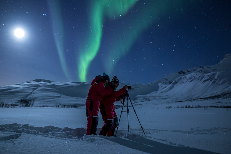 Two people in red snowsuits stand in snowy terrain at night, adjusting a camera on a tripod. Bright green Tromso Northern Lights and a full moon glow in the starry sky above snow-covered mountains—perfect for Arctic adventures Norway.
