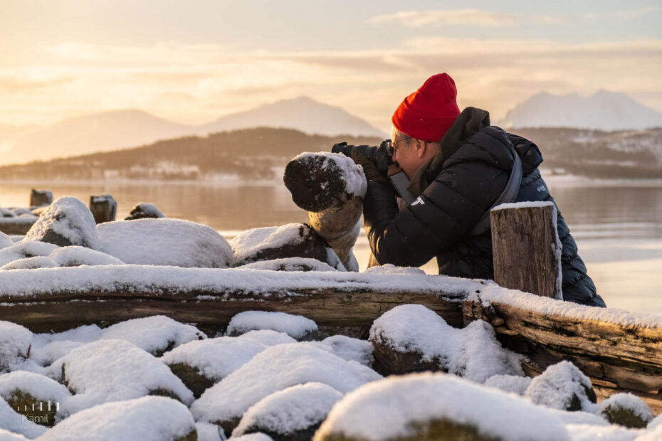 A person in a red beanie and dark jacket kneels on snow-covered rocks near a lake, using a camera to photograph the winter landscape with mountains in the background at sunrise or sunset.